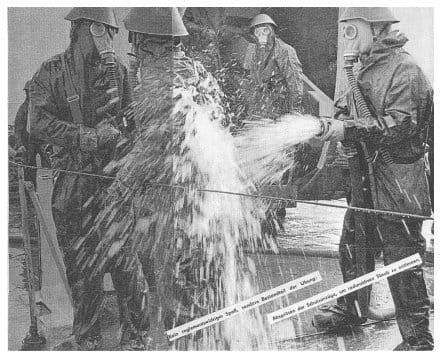 Zeitschriftenreproduktion: Vier Männer in Schutzanzügen und Gasmasken bei der Entaktivierung, einer spritzt mit einem Schlauch Wasser auf die Schutzkleidung der anderen