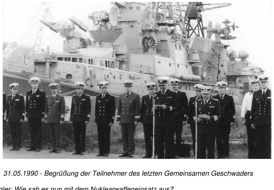 Eine Reihe von Marineoffizieren steht vor zwei am Kai liegenden Kriegsschiffen, in der Mitte spricht ein Offizier ins Mikrofon
