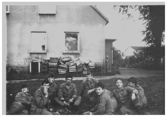 Acht Marineangehörige in Arbeitsuniform sitzen und liegen im Gras vor einem Dorfhaus, an dessen Wand leere Getränkekästen gestapelt sind